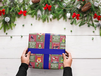 Das Bild zeigt eine Person, die ein in humorvollem Weihnachtspapier verpacktes Geschenk mit blauer Schleife hält. Im Hintergrund sind Tannenzweige, rote Schleifen, Kiefernzapfen und silberne Weihnachtsdekoration zu sehen.