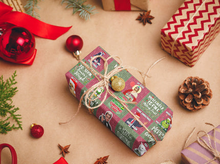 Das Bild zeigt ein weihnachtlich verpacktes Geschenk mit lustigen Motiven, darunter bekannte Figuren und Weihnachtsbotschaften. Es ist mit Juteschnur und einer goldenen Kugel dekoriert, umgeben von weiteren Geschenken und