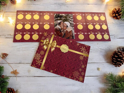 Ein Adventskalender in Rot mit 24 goldenen Kreisen und einem Foto eines Paares in der Mitte. Umgeben von weihnachtlicher Deko liegt der Kalender auf einem weißen Holztisch. Darunter ein roter Umschlag mit goldener Schleife und Schneeflocken.