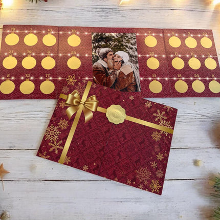 Ein Adventskalender in Rot mit 24 goldenen Kreisen und einem Foto eines Paares in der Mitte. Umgeben von weihnachtlicher Deko liegt der Kalender auf einem weißen Holztisch. Darunter ein roter Umschlag mit goldener Schleife und Schneeflocken.